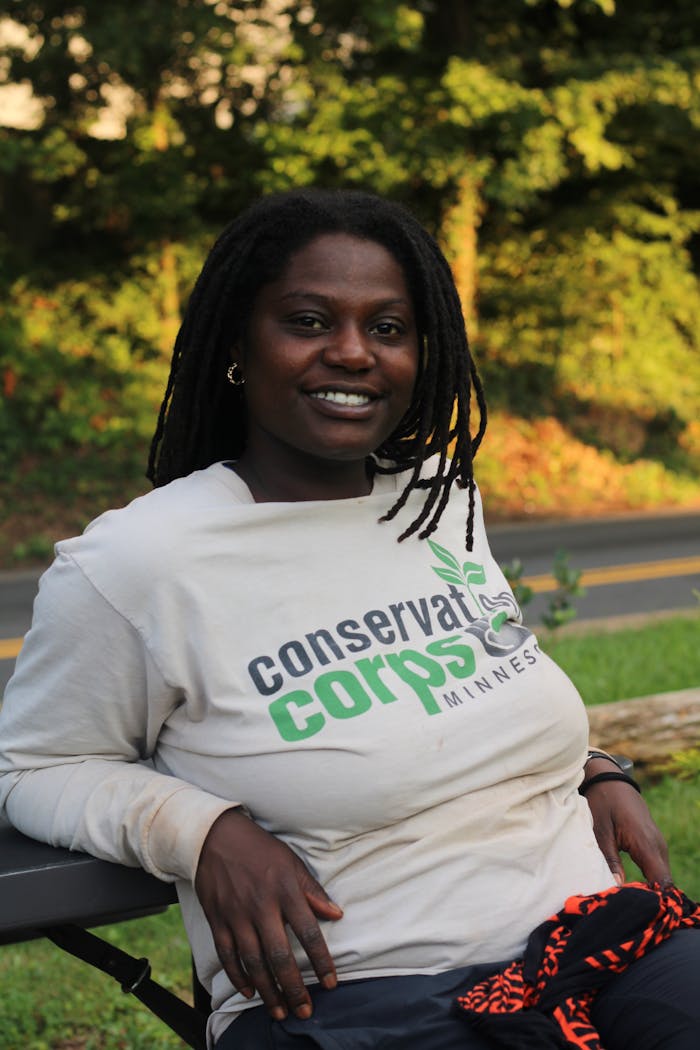 Smiling woman wearing a Conservation Corps shirt enjoys a sunny day outdoors.